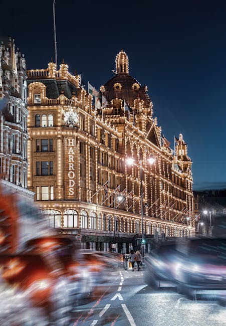 A nighttime exterior view of the Harrods department store in Knightsbridge, illuminated with warm, decorative lighting outlining its ornate architectural details and domed roof. The building features classical and Victorian-style stonework, large arched windows, and the 'Harrods' sign vertically displayed on the facade. The street below includes moving cars with blurred lights from their headlights and taillights, and a few pedestrians walking along the sidewalk. Bright streetlights cast a glow on the pavement, creating a vibrant urban scene that highlights the luxurious and historic ambiance of the Harrods building in Knightsbridge. For professional cleaning and maintenance of such historic and ornate buildings, Cleaners Knightsbridge offers expert services to maintain their shine and cleanliness aligned with top-quality standards.