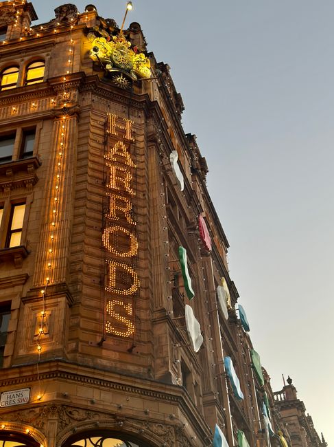 An exterior view of a historic multi-story building in Knightsbridge illuminated with warm lighting, featuring decorative architectural details and a large vertical sign spelling 'SALE ORDERS' in bright, illuminated letters. The building's facade showcases ornate stonework and large windows, some with laundry hanging outside, including colorful clothes and towels, indicating a lively urban setting. The scene is captured during dusk, with a clear sky and soft natural light highlighting the building's features. Cleaners Knightsbridge specializes in commercial cleaning, including surface cleaning, sanitisation, and deep cleaning services for buildings like this, ensuring hygiene and maintained appearance.
