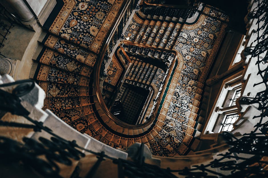 A high-angle view of a spiral staircase with ornate wooden handrails and decorative black iron balustrades. The staircase is lined with patterned, oriental-style carpets in shades of blue, cream, and brown, which cover each step and the landings. Sunlight streams through large windows on the right side, illuminating the wooden surfaces and highlighting the intricate details of the carpets. The design emphasizes the clean, polished condition of the surfaces, reflecting professional deep cleaning and maintenance by Cleaners Knightsbridge, specialists in surface cleaning for the Harrods area in Knightsbridge.