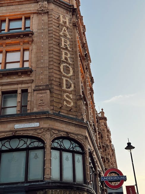 A nighttime view of the Harrods building in Knightsbridge illuminated with warm, decorative lights outlining its architectural features, including domes and turrets, with a clear dark sky in the background. The street in front shows blurred vehicles and pedestrians, highlighting the busy, lively atmosphere, while streetlights cast a gentle glow over the scene. Although the image focuses on the exterior of the ornate building, it exemplifies luxurious surroundings that benefit from professional cleaning and maintenance services offered by Cleaners Knightsbridge for their commercial and residential clients.