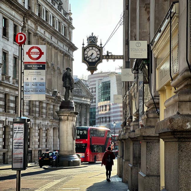A street scene in Knightsbridge with historic and modern buildings lining the sidewalk. There is a large, ornate clock mounted on a decorative metal bracket attached to a building, displaying the time as approximately 4:11. A statue of a man in formal attire stands on a pedestal near the corner, surrounded by stone pillars. A red double-decker bus is parked along the curb, and a person with a backpack is walking away from the camera on the sidewalk. The building facades feature detailed stonework and large windows. Street signage including a bus stop sign and a timetable are visible, along with several commercial signs hanging from the buildings, including one for 'TOMOKA'. The scene appears clean and well-maintained, capturing the timeless urban atmosphere of Knightsbridge as part of a professional cleaning service, with a focus on surface cleanliness and city maintenance, relevant to the Knightsbridge station short notice apartment cleaning service offered by Cleaners Knightsbridge.