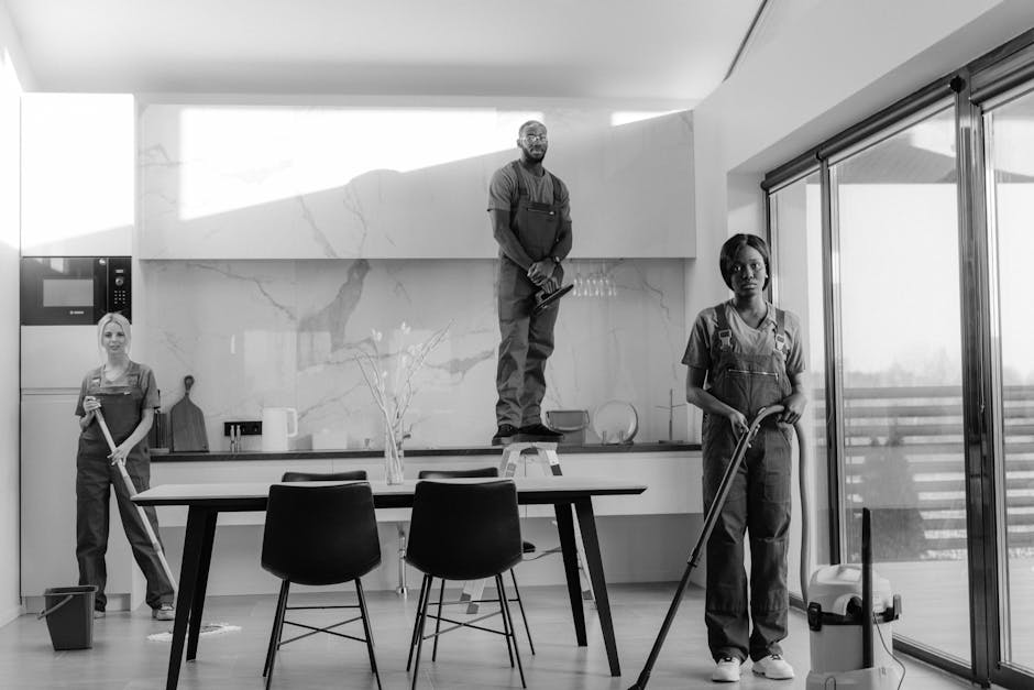 A black and white photo of three professional cleaners in a modern kitchen, each holding cleaning tools. One cleaner is standing on a step stool in front of a white marble backsplash, wearing work overalls and gloves. Another is standing near a sliding glass door with vertical blinds, holding a mop. The third cleaner is positioned near a black dining table, using a cleaning cloth on the surface. The kitchen features a stainless steel microwave, a small potted plant, and a utensil holder, with light pouring in through large windows. The scene emphasizes surface cleaning, deep cleaning, and sanitisation processes to maintain hygiene, as part of domestic cleaning services provided by Cleaners Knightsbridge.