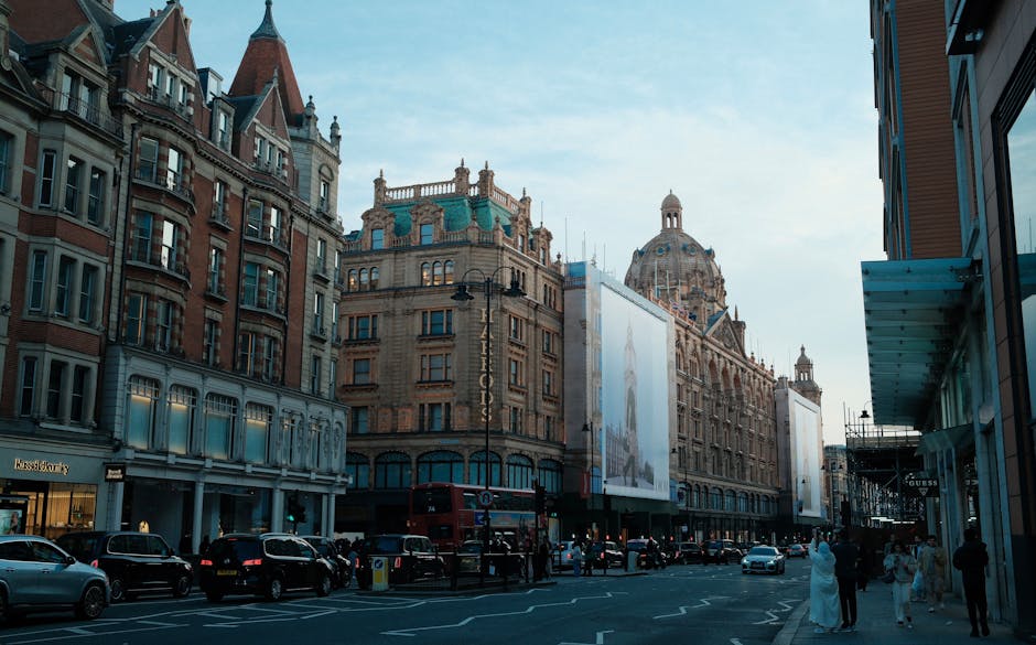 A street scene in Knightsbridge featuring historic and modern buildings with intricate architectural details, large commercial advertisements on the facades, and a line of parked cars along the curb. The street is bustling with pedestrians, some dressed in formal attire, walking on the sidewalk. The sky is partly cloudy with soft natural light illuminating the scene, highlighting the textures of the stone and glass surfaces. This image captures the vibrant urban atmosphere characteristic of Knightsbridge, with a mix of retail, office, and residential spaces, representative of the area served by Cleaners Knightsbridge for professional cleaning and maintenance services.